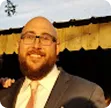 Smiling bald man with glasses and a beard wearing a suit and tie outdoors.